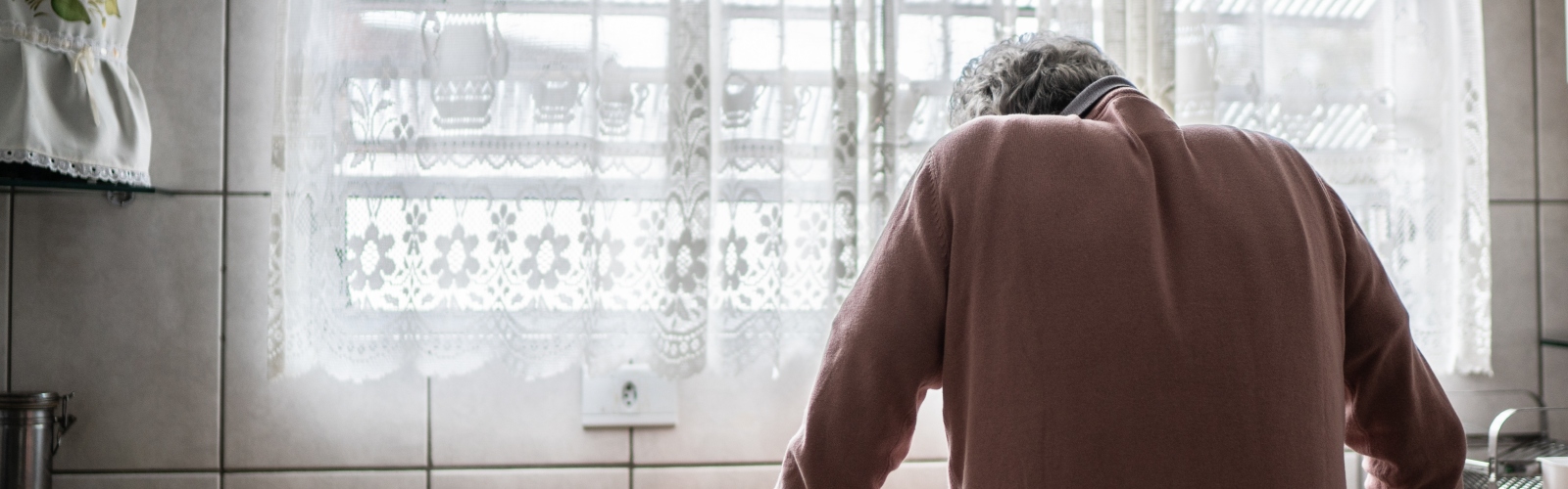 An older person in a pink jumper, standing in despair at a kitchen sink