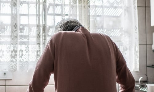 An older person in a pink jumper, standing in despair at a kitchen sink