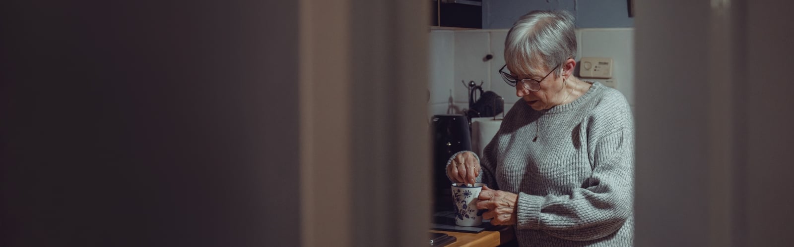 An older woman with short grey hair making a cup of tea, glimpsed through a slightly open door